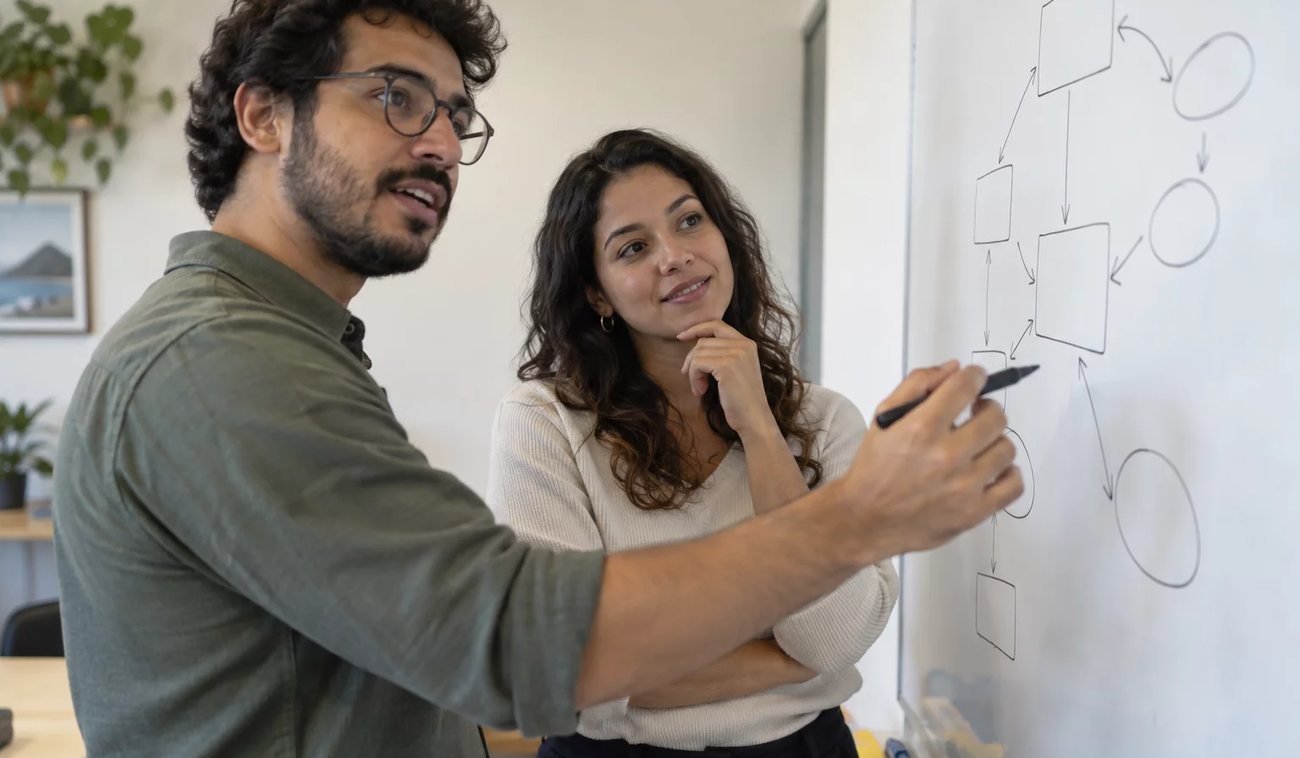 Três participantes conversam ao redor de uma mesa comprida numa sala clara de janelas altas